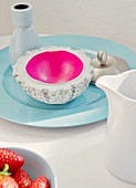 Concrete bowl with magenta inner surface and vase on pale blue porcelain plate, milk jug and bowl of strawberries in foreground