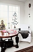 Round wooden table with turned legs painted black and white armchair in front of lattice windows in corner of wood-panelled room