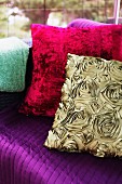 Kissen mit goldfarbenem Bezug in Seidenoptik und Rosenmuster mit rotem Plüschkissen auf lilafarbener Couch