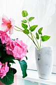 Weiß lasierte Vase mit pinkfarbener Blume, seitlich Zimmerpflanze mit rosa Blüten