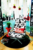 Swivel easy chair with black and white floral upholstery on round rug in corner; collection and black and red vases on windowsill