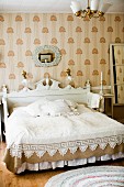 Double bed with white, carved headboard against wallpapered wall in traditional bedroom in shades of white and pale brown