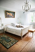 Simple bedroom with Regency-style double bed and bedside table painted white below chandelier wit glass pendants