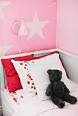 White, wooden child's bed with scatter cushions and teddy bear below wall lamp on pink wall with pattern of stars