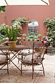 Delicate metal chairs and matching table on tiled floor of courtyard with potted plants against red-brown façade