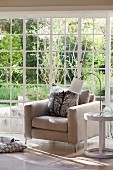 Modern armchair with pale upholstery, table lamp on side table and floor-to-ceiling lattice windows with view of garden