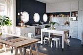 Table on castors in white fitted kitchen with black mosaic floor, black accent wall, porthole windows and wooden table on trestles in foreground