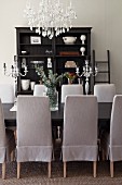Chairs with pale grey loose covers around dining table below chandelier; antique shelves in background