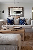 Ecru ottoman and sofa with scatter cushions around pale wooden coffee table below mirror with rustic wooden frame