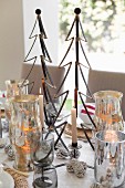 Brennende Kerze und Weihnachtsdekoration, stilisierter Tannenbaum aus Metall, zwischen Silber Windlichtern