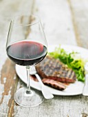 A glass of red wine in front of a grilled beef steak with salad