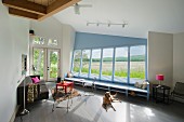 Minimalistische Loungeecke gegenüber gekippter, blau getönter Fensterfront mit Blick in die Landschaft, auf grauem Holzboden liegender Hund