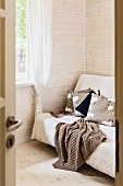 View through open door of blanket on sofa in corner of room with pale, varnished wooden walls