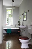 Toilet on glossy floor and vintage bathtub below window in background in bathroom painted pale grey