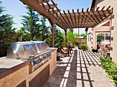 Aussengrill aus Edelstahl auf Terrasse teilweise mit Pergola aus Holz vor sonnenbeschienenem Landhaus