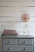 Dried allium seed head in glass vessel and stacked folded tea towels on grey-painted chest of drawers