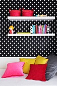 Decorative wall shelves on a wall hung with black and white spotted wallpaper above a sofa-bed with coloured cushions