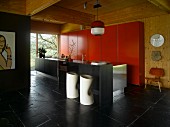 Zylindrisch geformte Barhocker mit Knick auf Schieferboden, vor schwarzer, freistehender Kücheninsel, dahinter roter Einbauschrank an Holzwand in offenem Wohnraum