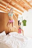 Junge Frau in Unterwäsche auf einem Bett in die Luft springend; im Hintergrund eine Frau bei der Kleiderauswahl