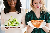 Two young women holding a plate of salad and a slice of pizza