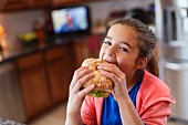 A girl in a kitchen biting into a large sandwich