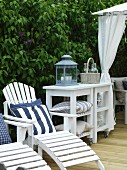 White-painted sun loungers with scatter cushions next to lantern on side table with castors on wooden deck