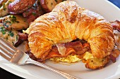 A croissant with scrambled eggs, bacon and cheese served with fried potatoes