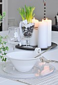 Place setting on dining table; bowl with handle decorated with cord, glass plate, wine glass and arrangement of candles and plants on tray