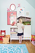 Girl standing in front of dolls' house on cabinet in child's bedroom decorated in bright colours