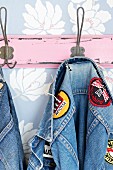 Denim jacket with fabric badges hanging on pink coat rack on pastel blue, floral wallpaper