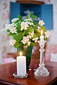 Stumpenkerze und Kerze in Glas Kerzenhalter vor Jasminblüten in grüner Vase auf Holztisch