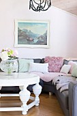 White coffee table in front of grey corner couch with scatter cushions below landscape painting on pastel pink wall