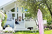 Sunny garden with seating area, woman sitting on steps leading to modern, white country house