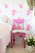 Pink-painted chairs with carved backrest and seat cushions against floral wallpaper next to pot of flowers on floor