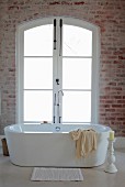 Bathmat in front of free-standing bathtub, white candle in large candlestick on floor and frosted French windows