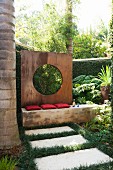 Stone flags amongst lawn in front of bench with red cushions and screen wall of rusty metal with circular aperture in tropical garden