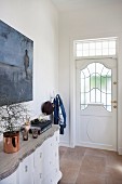 Chest of drawers with carved wooden top below picture on wall in foyer; front door with glass panels and transom light