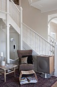 Armchair and magazine rack next to 50s-style speaker at foot of staircase with white-painted, wooden balustrade in foyer