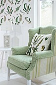 Armchair with pale green, patterned cover and scatter cushion next to white side table with table lamp; fern-patterned wallpaper