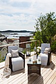 Wicker chairs with white cushions and wicker coffee table on wooden terrace with view of town and sea in background