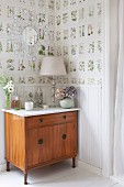 Tischleuchte auf Kommode in Zimmerecke, an Wand halbhohe, weiße Holzverkleidung und Tapete mit botanischem Muster