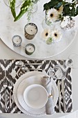 Place setting with white crockery and patterned table mat next to tealight holders and vases of flowers on round dish