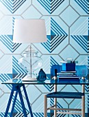 Tischlampe mit weißem Schirm auf Glastisch, vor Tapete mit geometrischem Muster in verschiedenen Blautönen