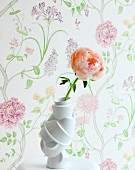 Apricotfarbene Rose in weisser Schnörkel Vase auf Ablage, vor Wand mit floral gemusterter Tapete in Pastelltönen