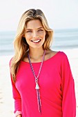 A young blonde woman on a beach wearing a pink sweatshirt and a long necklace