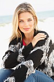 A young blonde woman sitting on a beach wearing a black and grey knitted cardigan