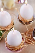 Egg-shaped candles in raffia nests in small dishes used as candle holders
