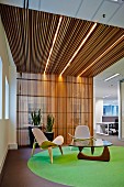 Klassiker Sessel mit hellen Polstern und Couchtisch auf grauem Teppich mit grünem Kreis im Foyerbereich, Wand aus vertikalen Holzlamellen in den Deckenbereich übergehend