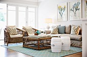 Lounge area with white, drum-shaped stools, wicker armchairs and sofa with scatter cushions around wicker coffee table and pastel blue, patterned rug; floral pictures on wall