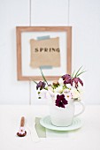 Kleiner Frühlingstrauss in weißem Tonkrug mit Stiefmütterchen, Primeln, Schachbrettblumen, Schneeball und Tazetten, im Hintergrund Holzrahmen mit Text an Wand